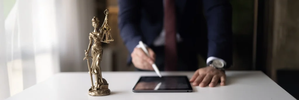 unscharfer Mann (Brust bis Gürtel sichtbar) steht hinter Tisch und schreibt auf Tablet, Justitia als Statue steht links auf dem Tisch